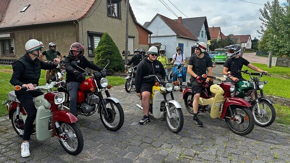 Junge Männer mit Motorradhelmen sitzen auf ihren Motorrädern vor einem Einfamilienhaus
