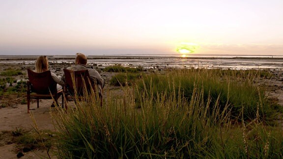 Eine Frau und ein Mann sitzen in Stühlen am Meer und genießen den Sonnenuntergang.