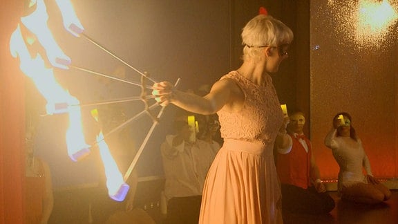 Eine Frau tanzt und hält dabei eine Vorrichtung mit fünf Fackeln in ihrer rechten Hand.