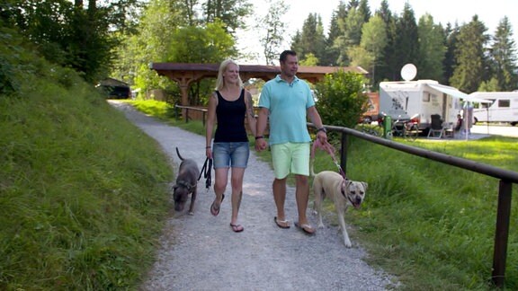 Eine Frau und ein Mann gehen mit zwei Hunden Hand in Hand spazieren, im Hintergrund sieht man den Campingplatz mit Wohnmobilen.
