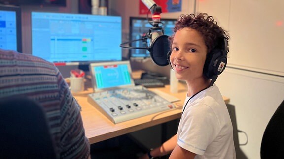 Johann, neun Jahre alt, sitzt im MDR Tweens Studio vor dem Mikro. Auf dem Kopf trägt er Kopfhörer, er lacht in die Kamera. 