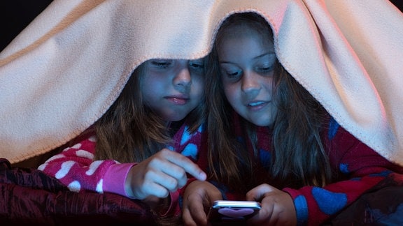 Zwei Mädels verstecken sich unter einer Decke und schauen auf ein Handy-Display. Sie tragen Pyjamas.