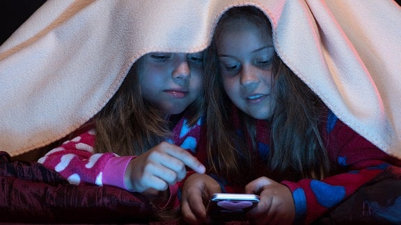 Zwei Mädels verstecken sich unter einer Decke und schauen auf ein Handy-Display. Sie tragen Pyjamas.