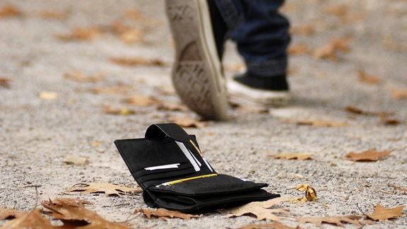Jemand geht über einen mit Herbstlaub bedeckten Weg. Hinter ihm liegt auf dem Weg eine verlorene Brieftasche. 