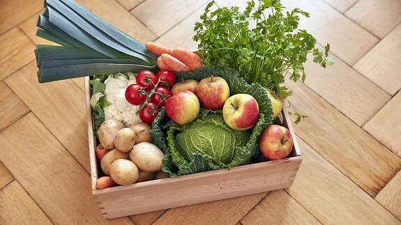 Auf einem Parkettfußboden steht eine Holzkiste mit Obst und Gemüse