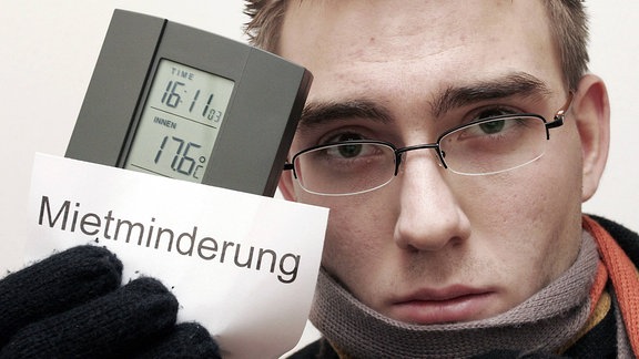 Ein junger, warm angezogener Mann, hält in seiner Hand ein Thermometer, das eine Innentemperatur von 17,6 Grad anzeigt, sowie einen Zettel mit der Aufschrift "Mietminderung"