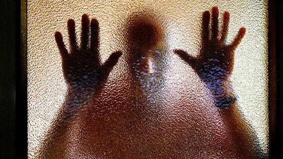 An einer Glastür sind die Umrisse eines Mannes zu sehen, der von der anderen Seite seinen Kopf und beide Hände an die strukturierte Glasscheibe hält.