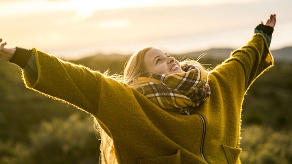 Eine junge Frau streckt draußen lachend ihre Arme aus