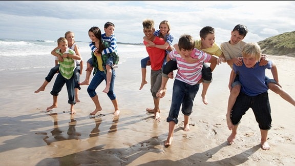 Jugendliche rennen, sich huckepack tragend, einen Strand entlang.