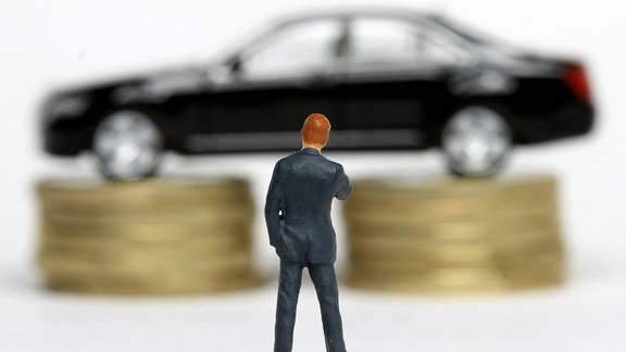 Mann im Anzug steht vor einer auf einem Stapel Euro Geldmünzen aufgebockten dunklen Luxuslimousine
