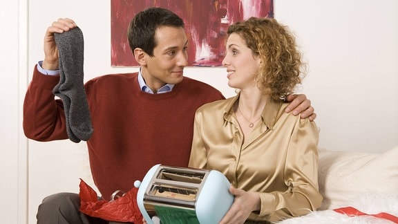 Eine Frau und ein Mann sitzen mit ihren Weihnachtsgeschenken auf dem Sofa und spielen sich Freude vor. Er hält ein paar graue Socken hoch, sie hält auf ihrem Schoß einen Toaster.