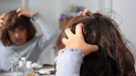 Ein kleines Mädchen steht vor dem Spiegel und kratzt sich mit beiden Händen am Kopf.