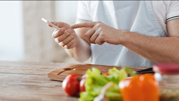 Ein Mann mit Schürze sitzt an einem Küchentisch und bedient ein Smartphone, vor Ihm auf dem Tisch liegt Gemüse