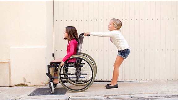 Ein Mädchen schiebt ein anderes Mädchen, das im Rollstuhl sitzt.