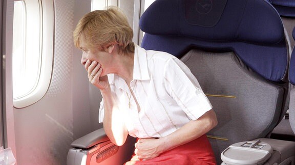 Frau schaut während eines Fluges aus dem Fenster und hält dabei eine Hand an den Bauch und die andere vor den Mund