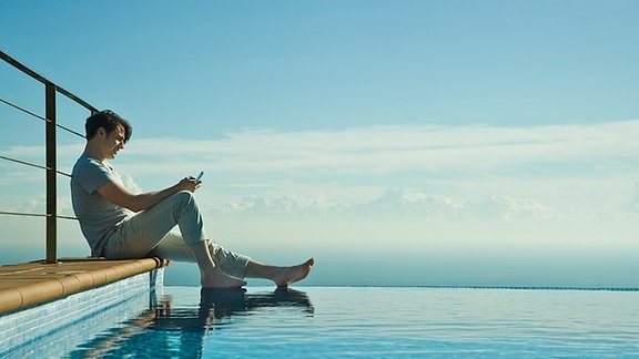 Ein Mann sitzt mit einem Mobiltelefon an einem Pool