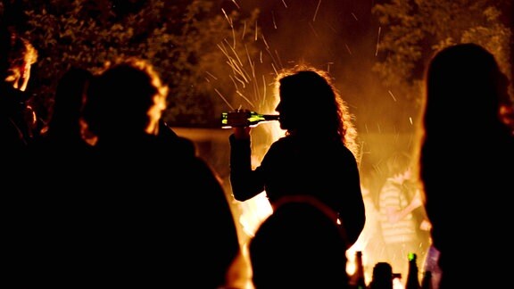 Eine Nachtaufnahme zeigt den Umriss einer jungen Frau, die während einer Party zwischen anderen an einem Lagerfeuer steht und Bier aus der Flasche trinkt