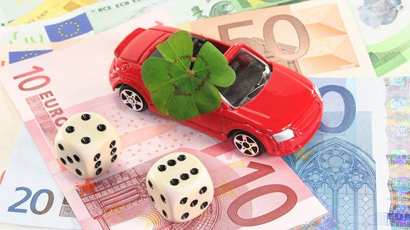 Ein rotes Spielzeugauto mit einem vierblättrigen Kleeblatt steht auf Geldscheinen, daneben zwei Würfel.