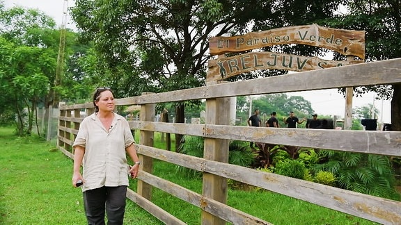 Eine Frau geht an einem Zaun entlang, im Hintergrund steht eine Gruppe Menschen.