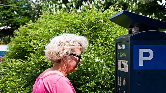 Seniorin mit Sonnenbrille vor einem Parkscheinautomaten