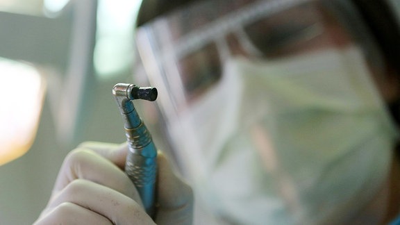 Ein Zahnarzt mit Schutzvisier beugt sich ein Poliergerät in der Hand haltend über einen Patienten. 