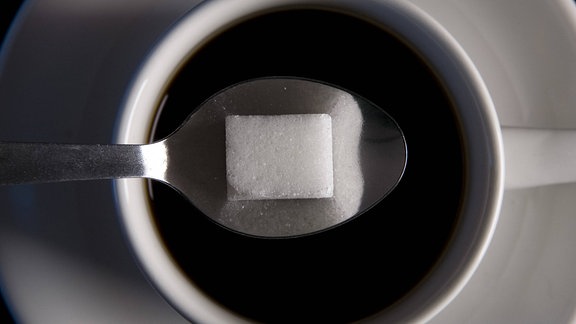 Draufsicht auf ein Stück Würfelzucker, das auf einem Teelöffel liegt, der sich über einer Tasse Kaffee befindet. 