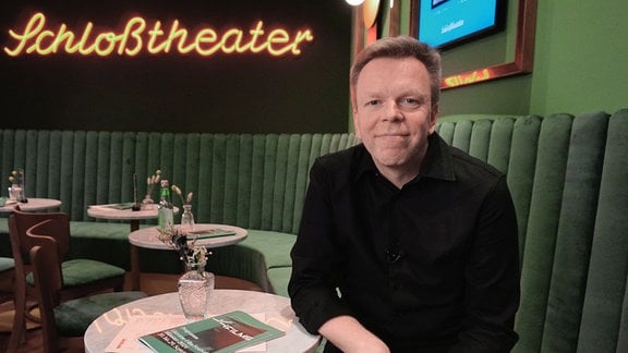 Eine männlcihe Person sitzt auf einem grünen Sofa. Dahinter der LED-Schriftzug Schlosstheater. Portraitfoto von Carsten Happe.