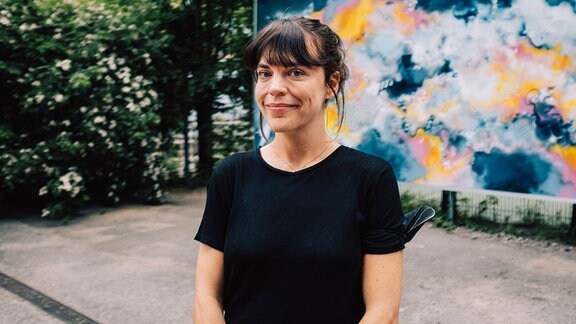 Porträt der Leiterin des Mo&Friese - Junges Kurzfilm Festival Hamburg, Lina Paulsen. Eine junge Frau mit mittellangen dunklen Haaren und einem schwarzen Oberteil mit kurzen Ärmeln. Sie steht vor einer bunt bemalten Wand in einem begrünten Hinterhof. 