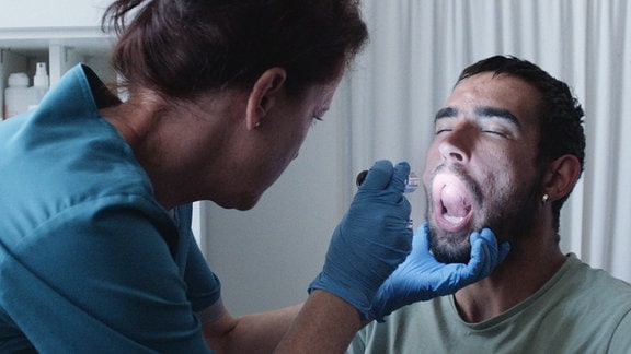 Ein Mann befindet sich in einem klinischen Setting und hat den Mund weit geöffnet. Eine Frau in blauer Kleidung und mit blauen Einweghandschuhen leuchtet ihm mit einer Taschenlampe in den Mund.