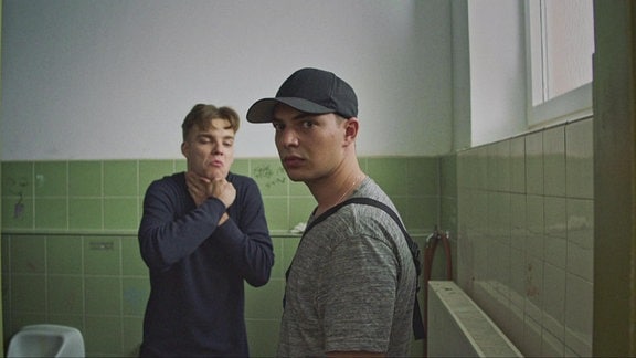 Zwei junge Männer sind auf einer Herrentoilette, einer der beiden hat die Hände um seinen Hals geklammert