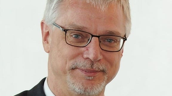 Mann mit kurzen grauen Haaren, kurzem grauen Bart und schwarzer Brille schaut in die Kamera