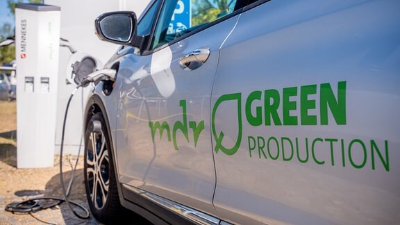 Ein weißes E-Auto steht hinter einer Ladesäule, auf der "MDR green Production" steht