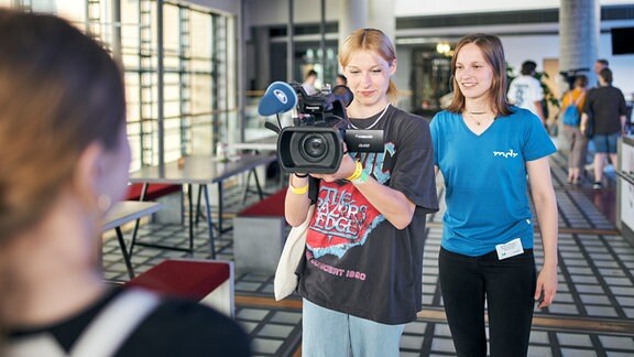 Eine junge Frau erklärt einer zweiten jungen Frau, den Umgang mit einer Videokamera, die sie in der Hand hält.