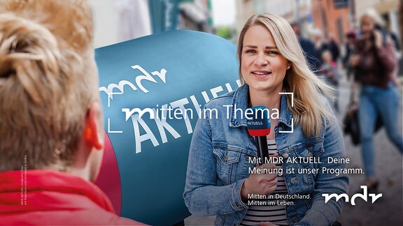 Eine junge blonde Frau sitzt auf einem Sofa und hält ein Mikrofon in der Hand. Sie interviewt eine zweite Person mit einer roten Jacke. 