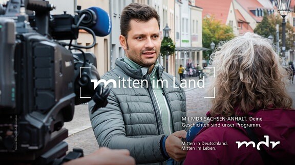 Ein Mann interviewt eine Frau mit einem Mikrofon. Beide stehen in einer Fußgängerpassage einer Altstadt. Im Vordergrund ist eine Fernsehkamera zu sehen. 
