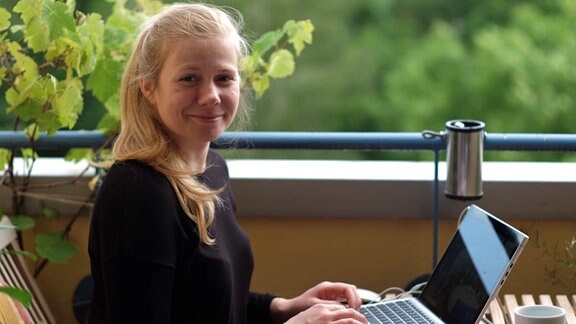 Luise Schönwald sitzt auf einem Balkon mit ihrem Laptop und lächelt in die Kamera