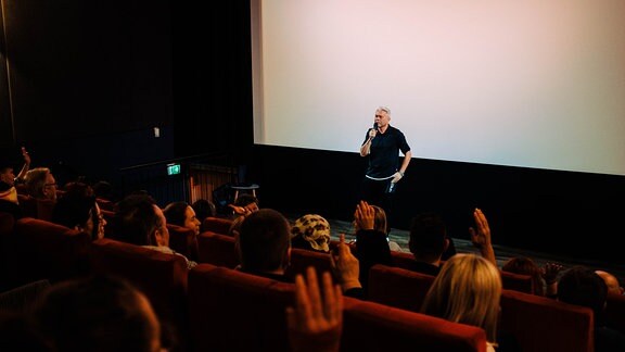 Mann auf der Bühne spricht mit Mikrofon zu Publikum im Kinosaal