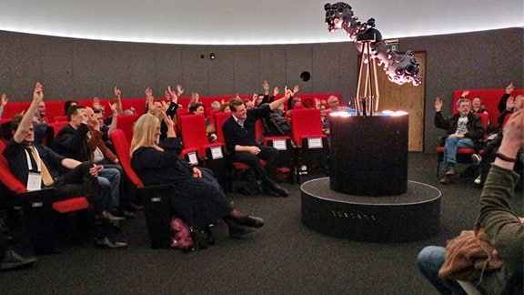 Mehrere Menschen sitzen in einem Planetarium im Kreis auf roten Sesseln.