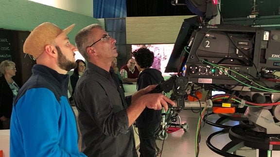 Zwei Männer blicken in einem Studio auf eine TV-Kamera.
