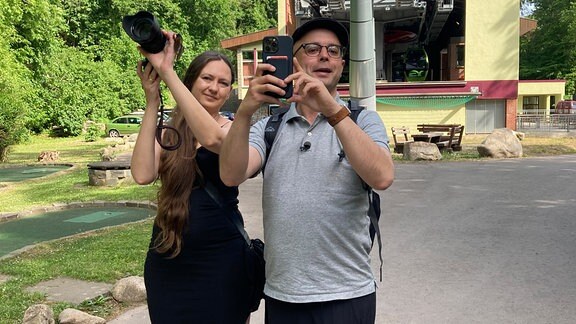 Mann und Frau stehen mit Fotokameras vor Seilbahn.