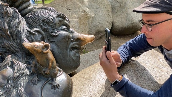 Mann hockt auf Felsen und fotografiert eine Hexen-Figur mit dem Smartphone.