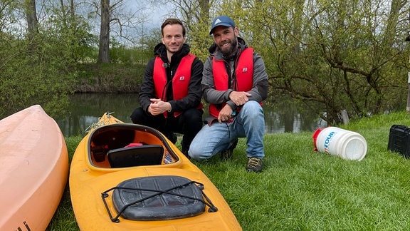 Zwei Männer posieren neben einem Boot am Ufer.