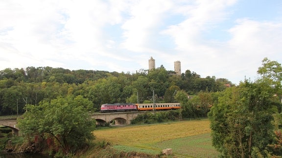 Ein Zug mit einer roten Lok fährt unterhalb einer Burgruine über eine Brücke. 