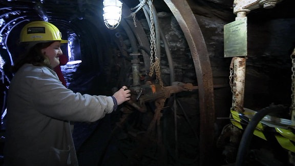 Frau mit Helm und weißen Kittel an einem Bohrer in einem Bergbaustollen