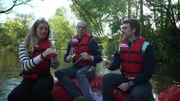 Drei Personen auf einem Paddelboot prosten sich mit Wein zu.