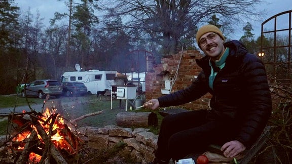 Mann sitzt am Lagerfeuer auf einem Campingplatz im Hintergrund stehen Wohnmobile