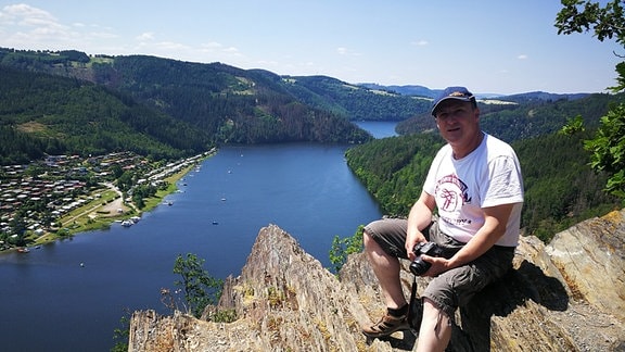 Mann mit Kamera sitzt auf einem Felsen im Hintergrund das Thüringer Meer