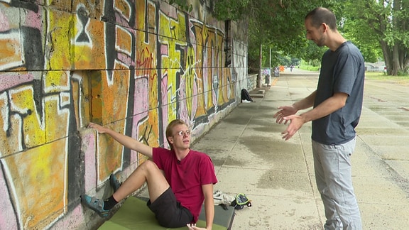 Der Volontär Lukas sitzt neben einer Mauer mit den Händen an der Wand. Daneben steht Michael, der ihm zeigt, wie man bouldert.