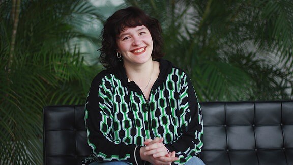 Volontärin Corinna Thamm sitzt lächelnd auf einem Lederstuhl vor Palmen. 