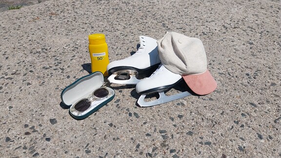 Ein Paar Schlittschuhe liegen in der Sonne auf dem Boden. Daneben steht eine Flasche Sonnencreme, eine Sonnenbrille und ein Basecap.